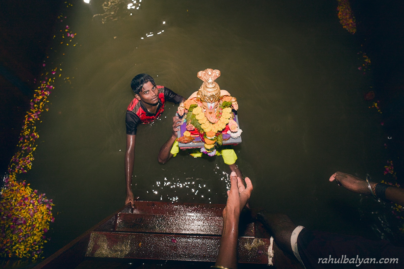 Top-down view of a young man in dark water receiving a small Ganesha idol from outstretched hands, flower petals floating