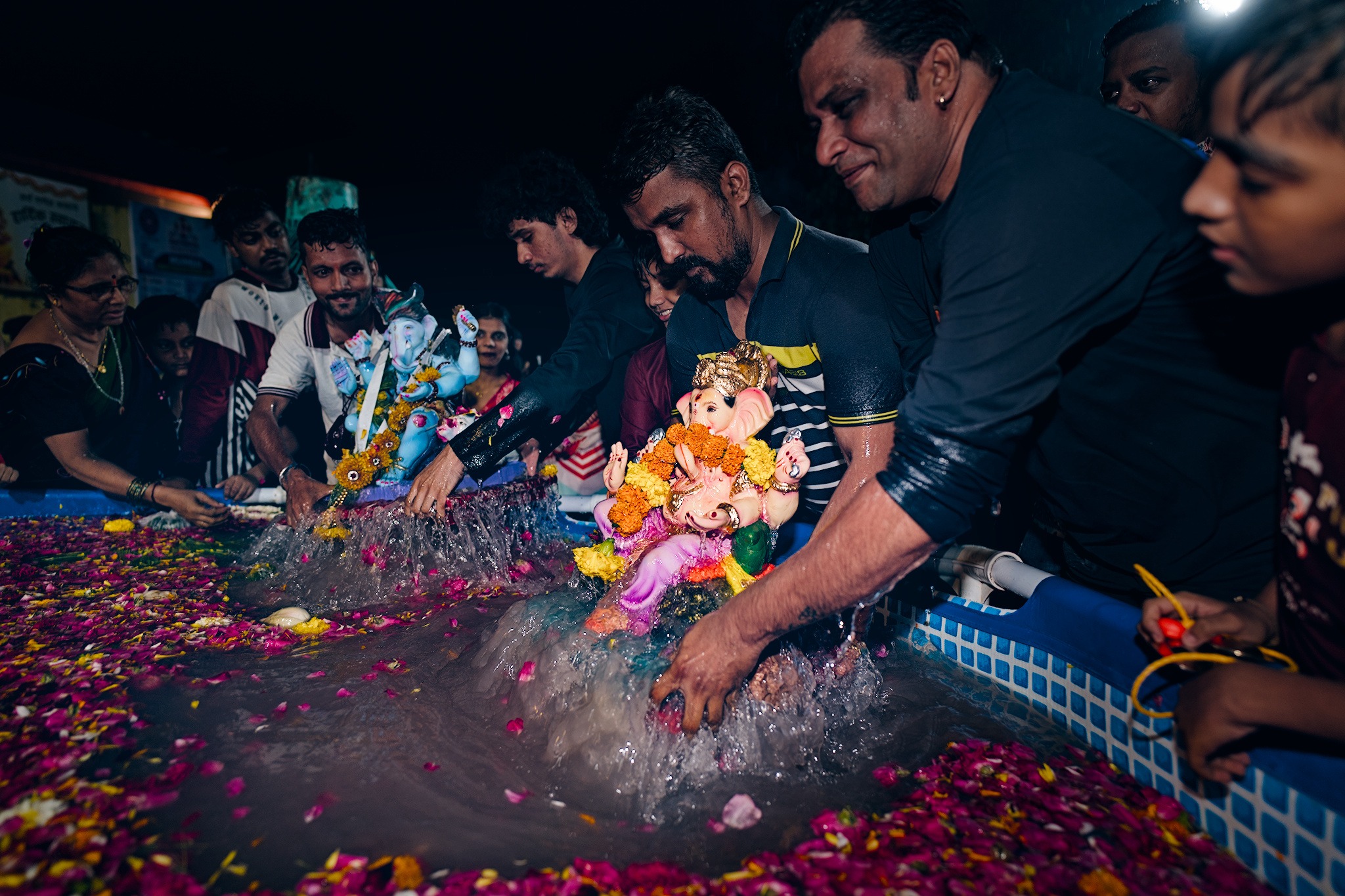 Men immerse small Ganesha idols into a flower-strewn artificial pond at night, water splashing