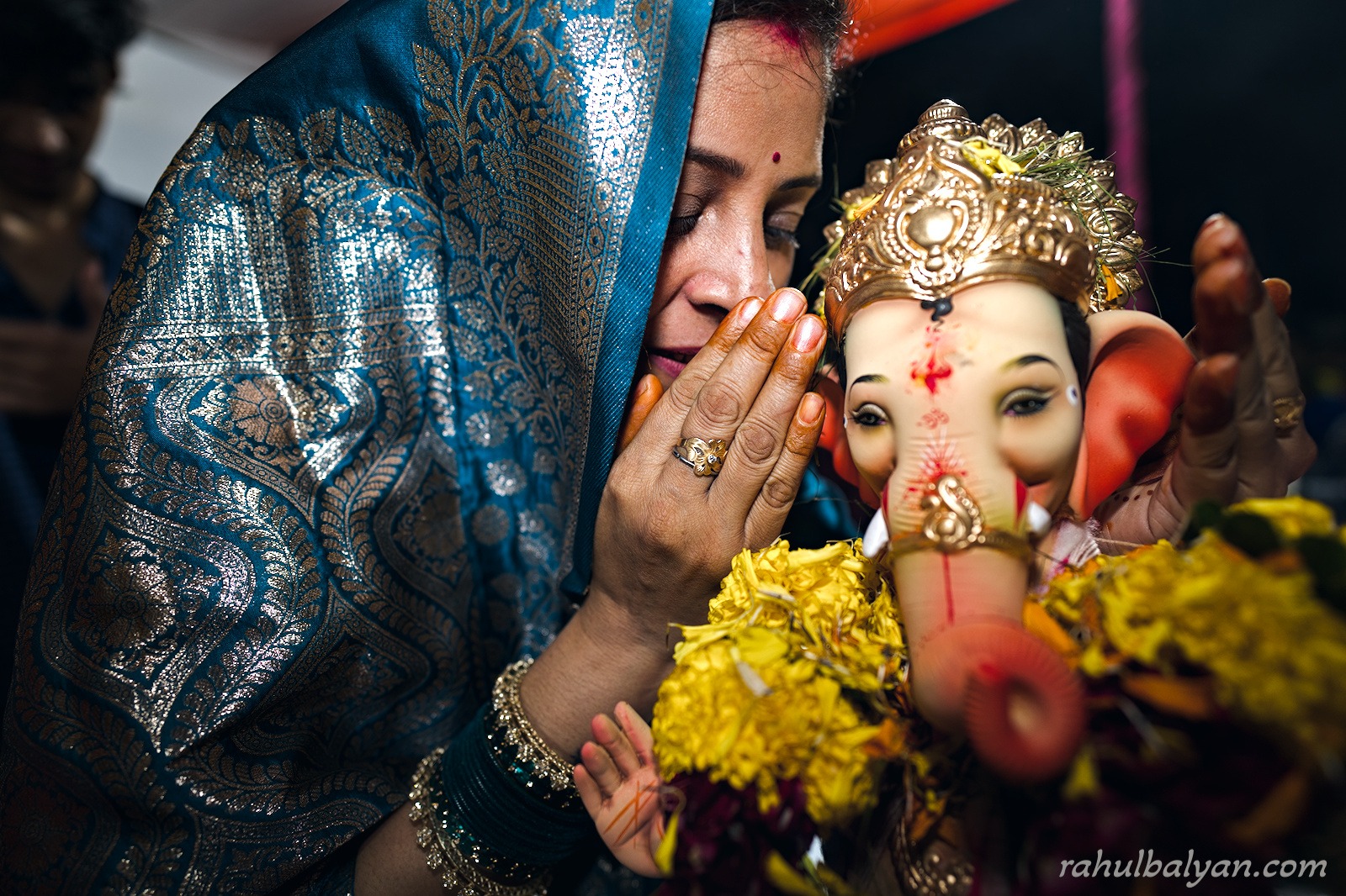 Woman in a teal-and-silver saree bows her head in prayer beside a small Ganesha idol with marigolds