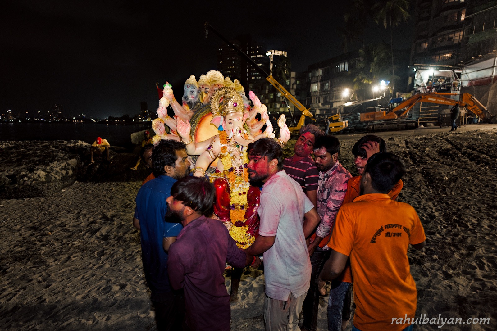 Group of men with gulal-smeared faces carry a multi-armed Ganesha idol across a beach at night, cranes and towers behind