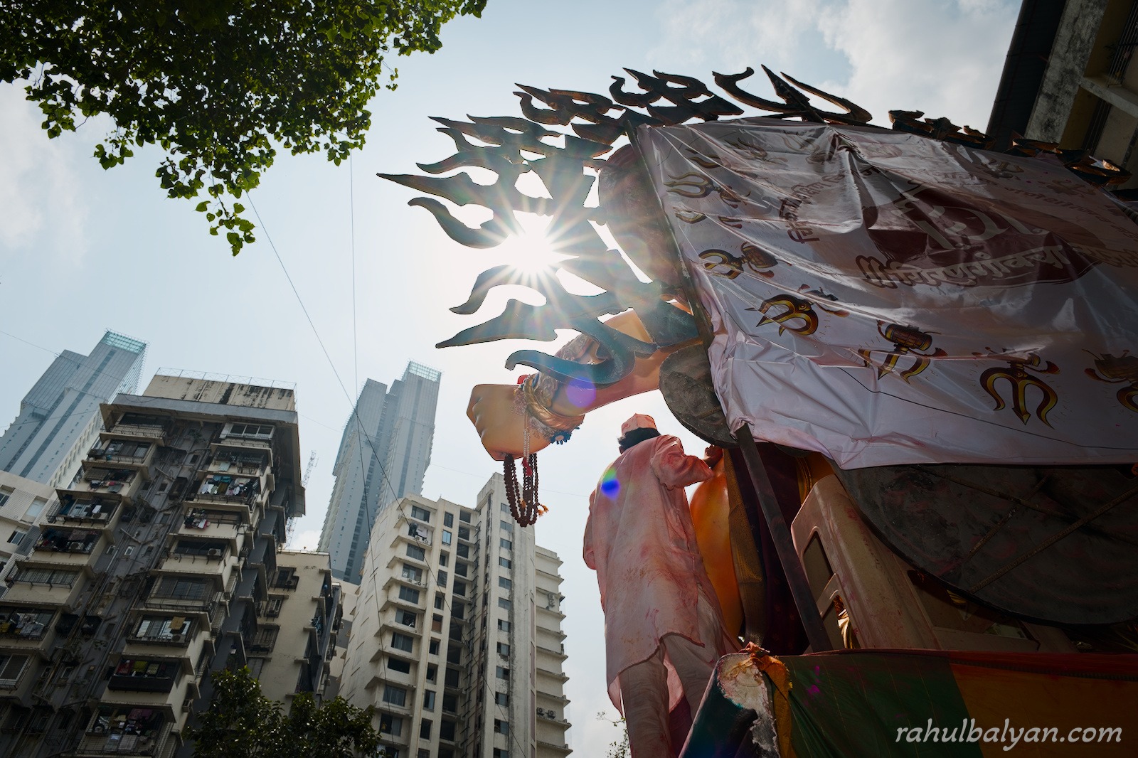 Low-angle view of a giant idol's spiked halo with the sun bursting through, Mumbai high-rises behind
