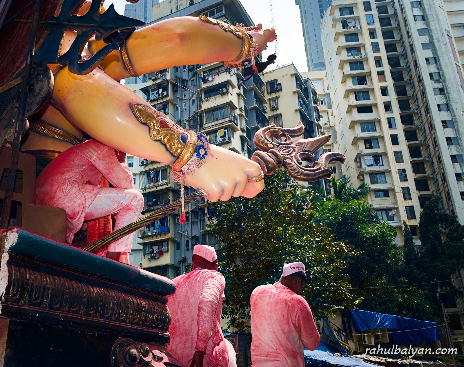 Giant idol's arm holding a trishul reaches over two men in pink kurtas, apartment towers behind