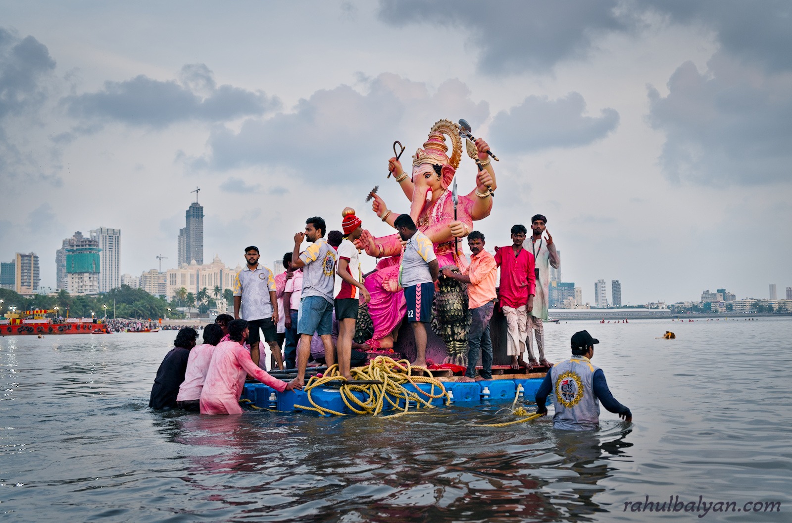 Lifeguards in yellow shirts lift a Ganesha idol on a fish-shaped float at night, city lights behind