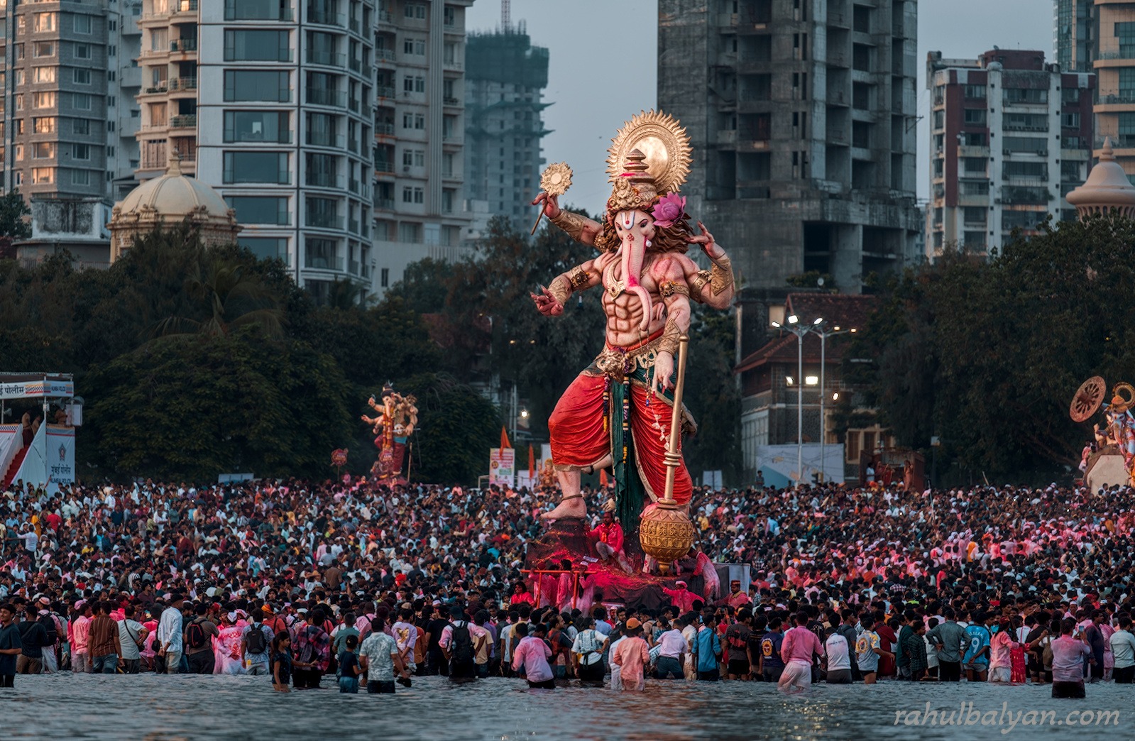 Tall Ganesha idol carried through waist-deep water by a vast crowd at dusk, high-rises behind