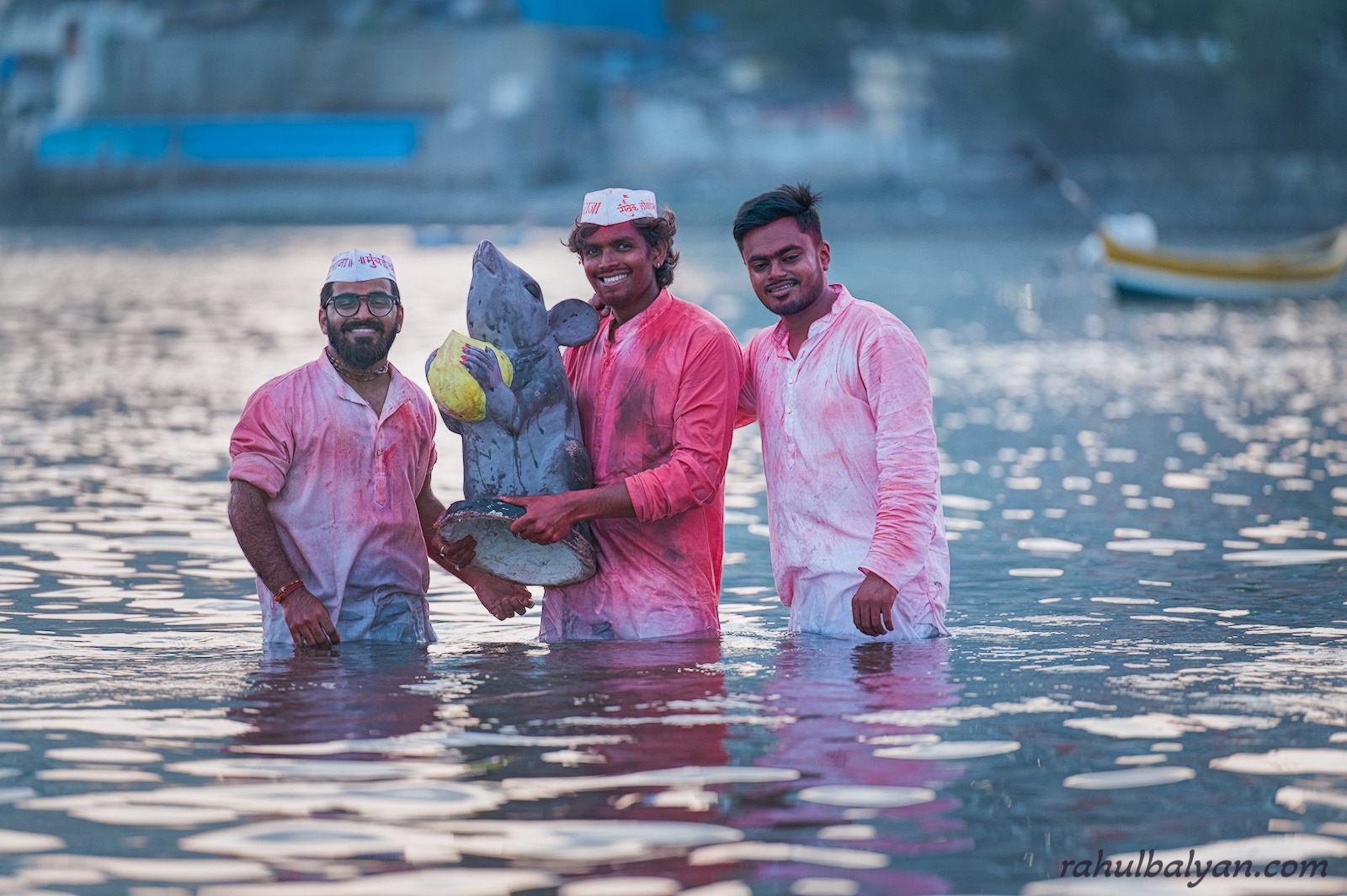 Three men in pink-stained kurtas stand smiling in the sea holding an idol of Mooshak, Ganesha's mouse